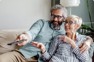 A middle-aged man and woman lounge on a couch together. The man points a remote control.