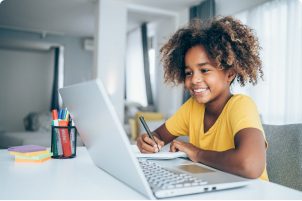 A girl around 10 years old looks at a laptop on her desk and is taking notes with pen and paper.