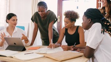 Four women are collaborating at a table. They are using pen and paper and an iPad.
