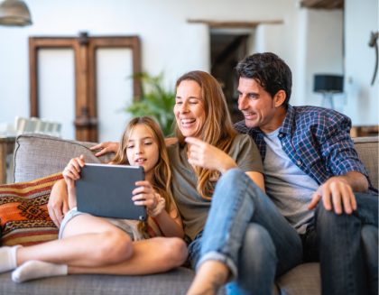 A mother, father and daughter are sitting on a couch. They are smiling and watching the tablet that the daughter is holding.