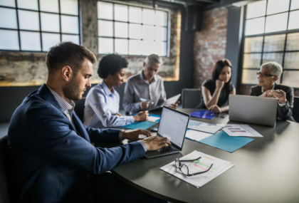 A meeting is taking place in an office room amongst five employees. Two have open laptops and one person is taking notes on paper.