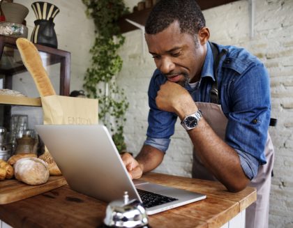 A baker wearing an apron uses a laptop at his place of employment.