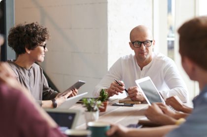 Employees sitting at a table in a well-lit business setting. Many have laptops and tablets.