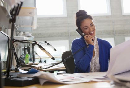 A woman is smiling while talking on the phone in a well-lit office. She holds large printouts in her other hand.