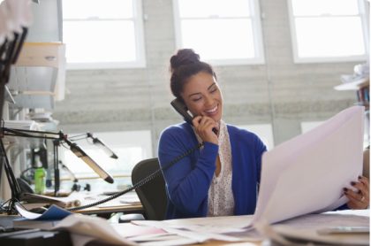 A woman is smiling while talking on the phone in a well-lit office. She holds large printouts in her other hand.