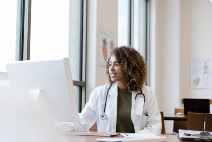 A doctor sits at an office desk next to a sunlit window. She is using a desktop computer and smiling. A stethoscope hangs around her neck.