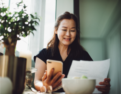 A woman sits at a table, smiling while she looks at a smartphone and a paper invoice.