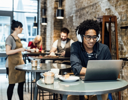 Man works on a laptop at a coffee shop table. A waitress and another patron are in the background having a conversation.