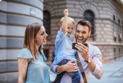 A mother, father and toddler-aged son are standing outside of a building on a sunny day. The father holds a smartphone for his son to view and they are all cheering.