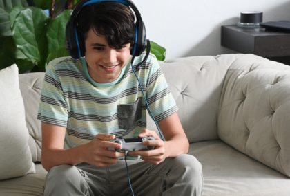 A teenage boy in a striped t-shirt sits on an off-white couch and plays a video game while wearing a large headset with a microphone.