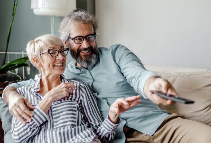A middle-aged man and woman lounge on a couch together. The man points a remote control.