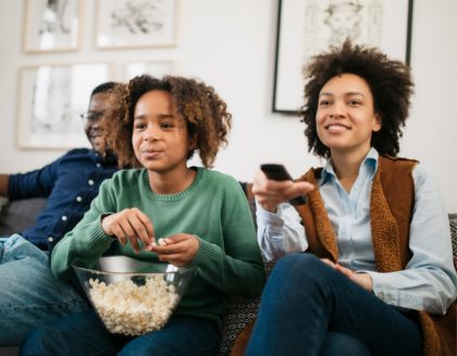A child eating from a bowl of popcorn sits with parents on a couch. The mother is pointing a remote control.