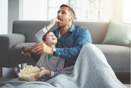 A father and son enjoying a movie while eating popcorn. The father is holding a remote control.
