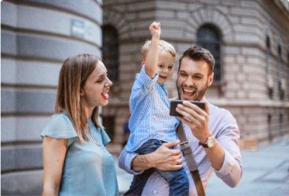 A mother, father and toddler-aged son are standing outside of a building on a sunny day. The father holds a smartphone for his son to view and they are all cheering.
