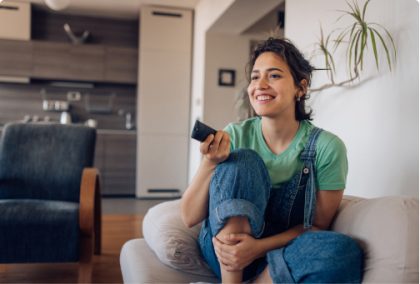 A woman in overalls sits comfortably with a remote control in her hand.
