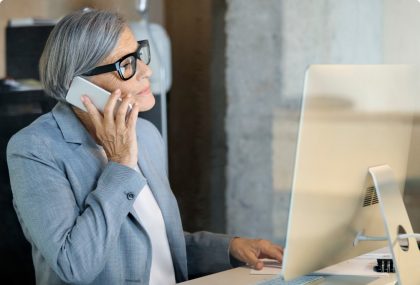 A professional woman speaking on a smartphone while also utilizing a desktop computer.
