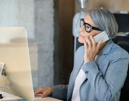 A professional woman speaking on a smartphone while also utilizing a desktop computer.