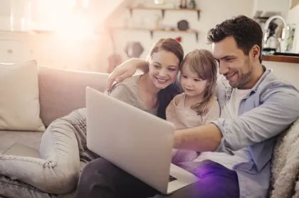 A smiling mother, father, and young daughter sit together on a couch looking at a laptop screen.
