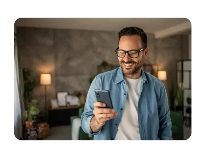 A smiling man with glasses and a blue button-down shirt looks at his smartphone in a modern, cozy living room.