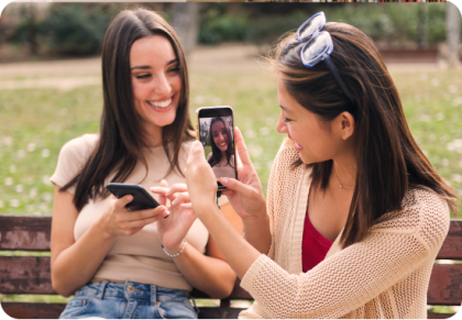 Two women sitting on park bench smiling and taking pictures with mobile phones