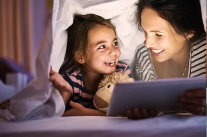 A mother and young daughter smile at each other while using a tablet under a white blanket fort.
