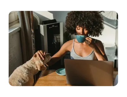 Woman sipping from mug at table with a laptop while petting dog