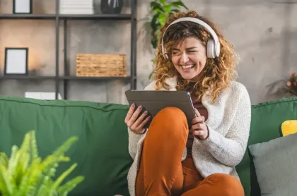 A woman with curly hair wearing headphones laughs while looking at a tablet on a green sofa.