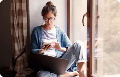 A woman sitting on her windowsill and typing on her laptop.