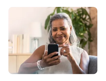 A smiling older woman with grey hair taps the screen of her smartphone while sitting on a couch at home.