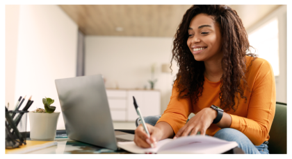 Student smiling and writing in notebook beside laptop